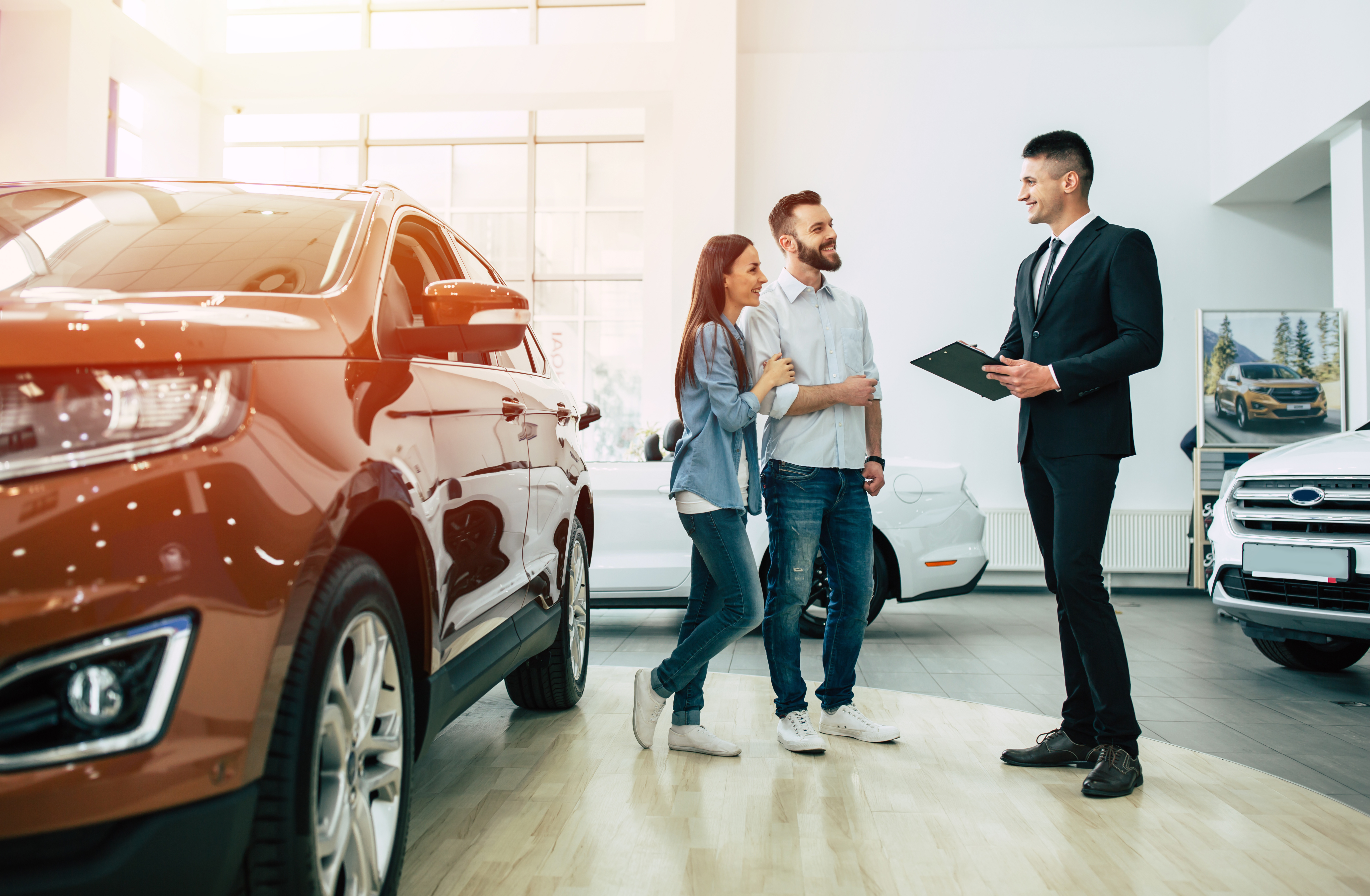 A smiling couple speaking with a professional salesperson holding a clipboard in a bright car showroom, with a shiny brown SUV in the foreground and other cars in the background.