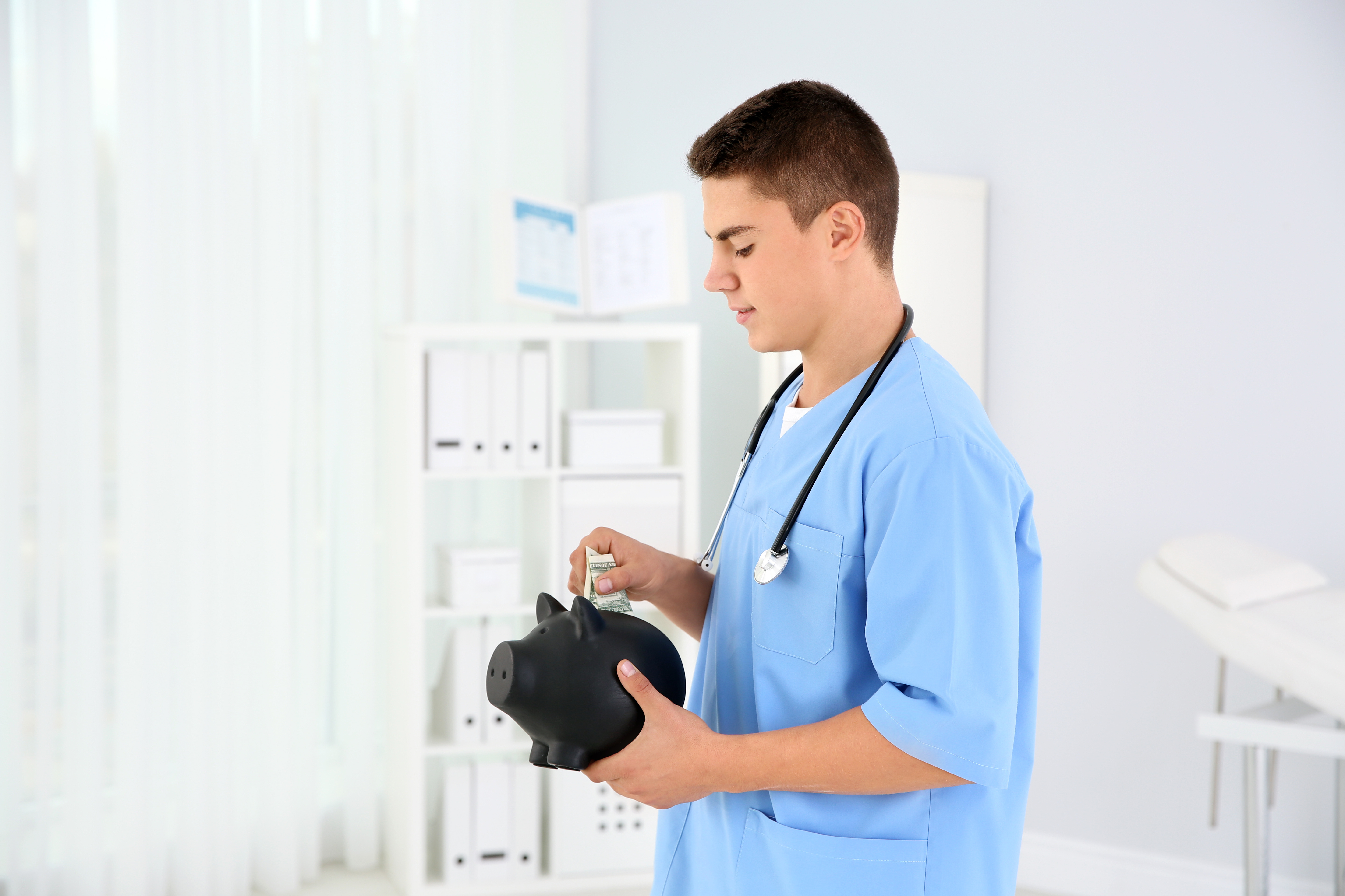 Young medical professional in blue scrubs holding a black piggy bank and inserting cash, symbolizing financial savings or planning for healthcare workers.