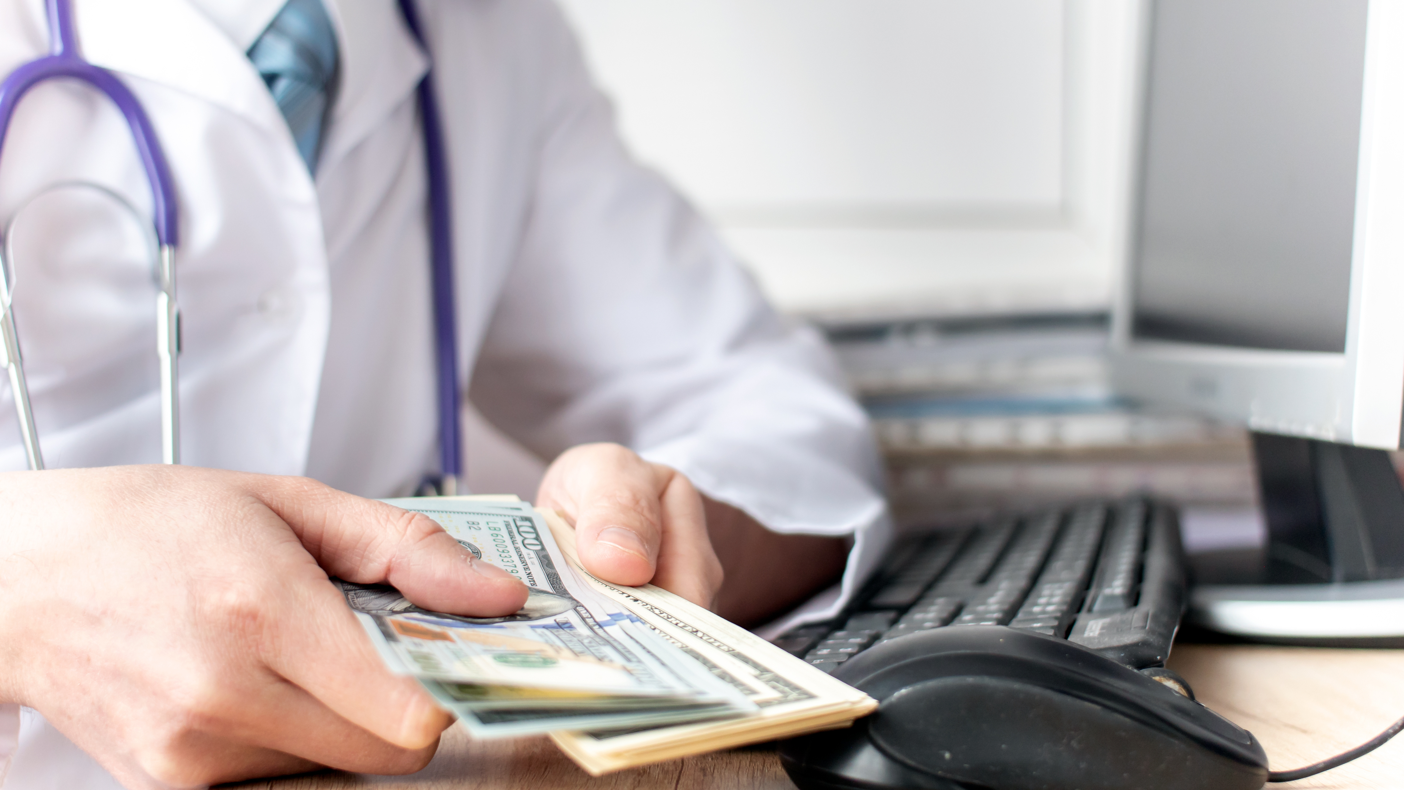 Close-up of a doctor holding cash while sitting at a computer desk, symbolizing financial management for healthcare professionals.