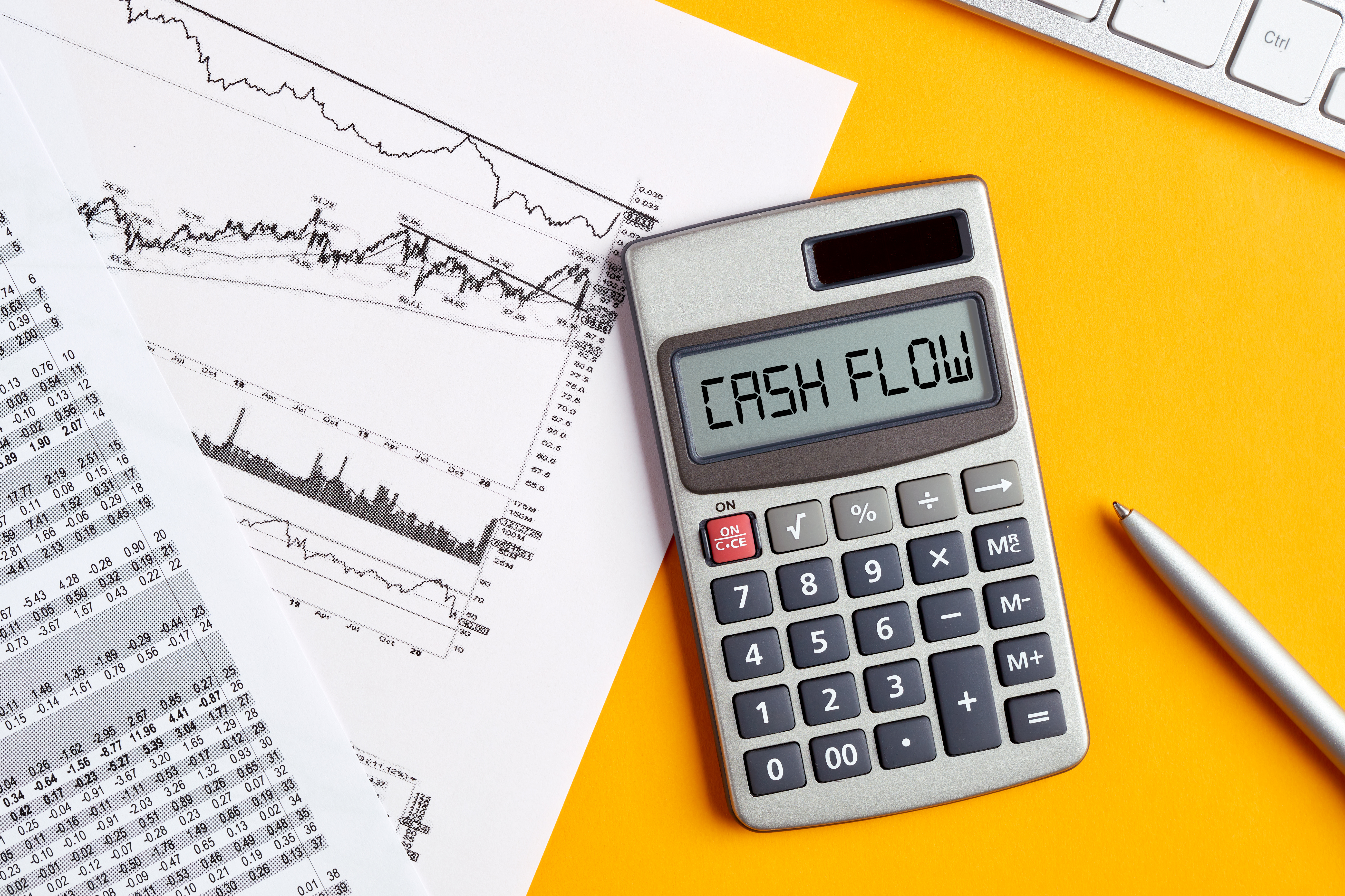 A calculator displaying 'Cash Flow' alongside financial graphs, spreadsheets, and a pen on a bright yellow background, symbolizing financial analysis and planning.