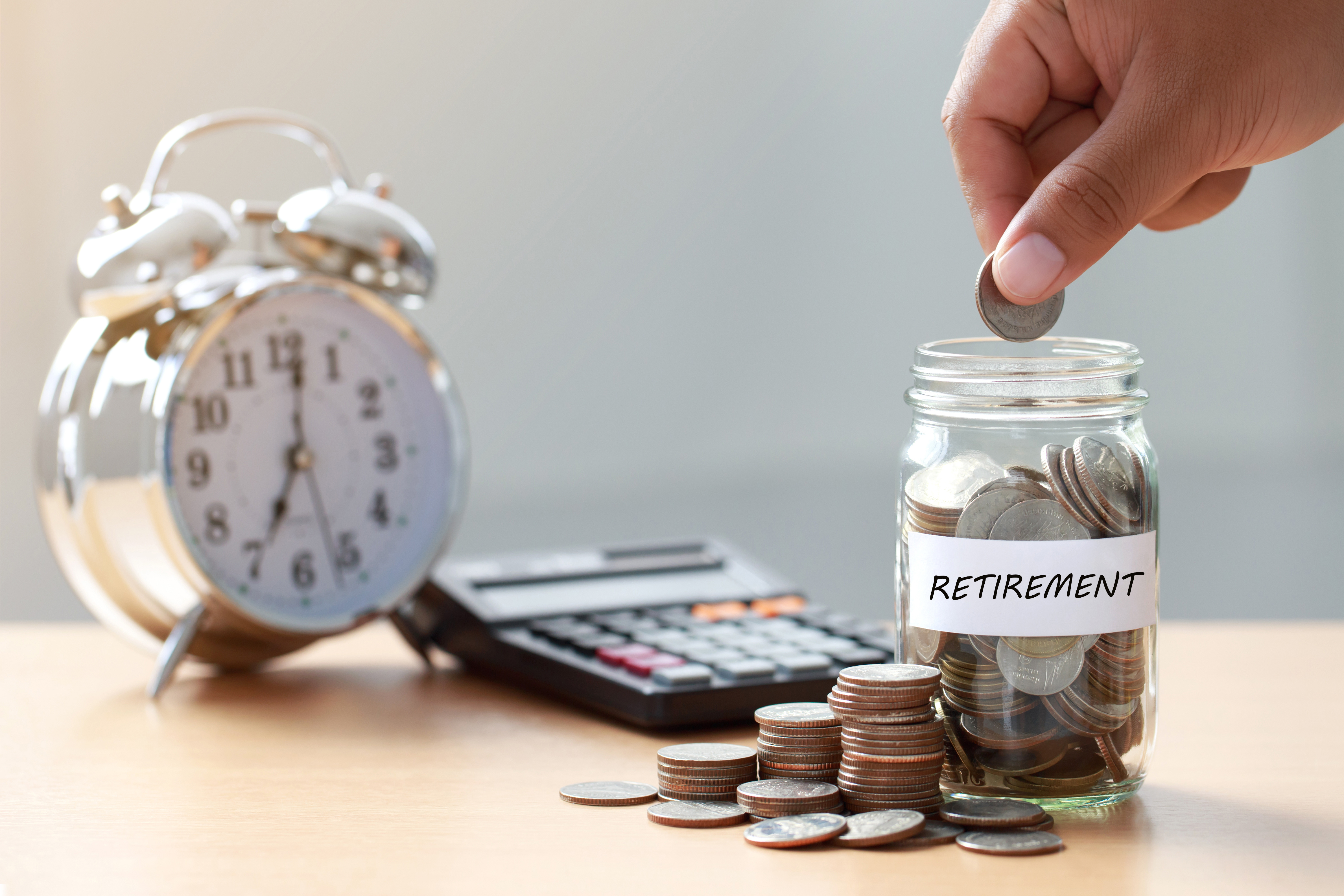 Hand puts a coin into a jar with the word retirement written on it. Right next to it is a clock symbolizing the passage of time and a calculator suggesting the need to calculate your retirement contributions.