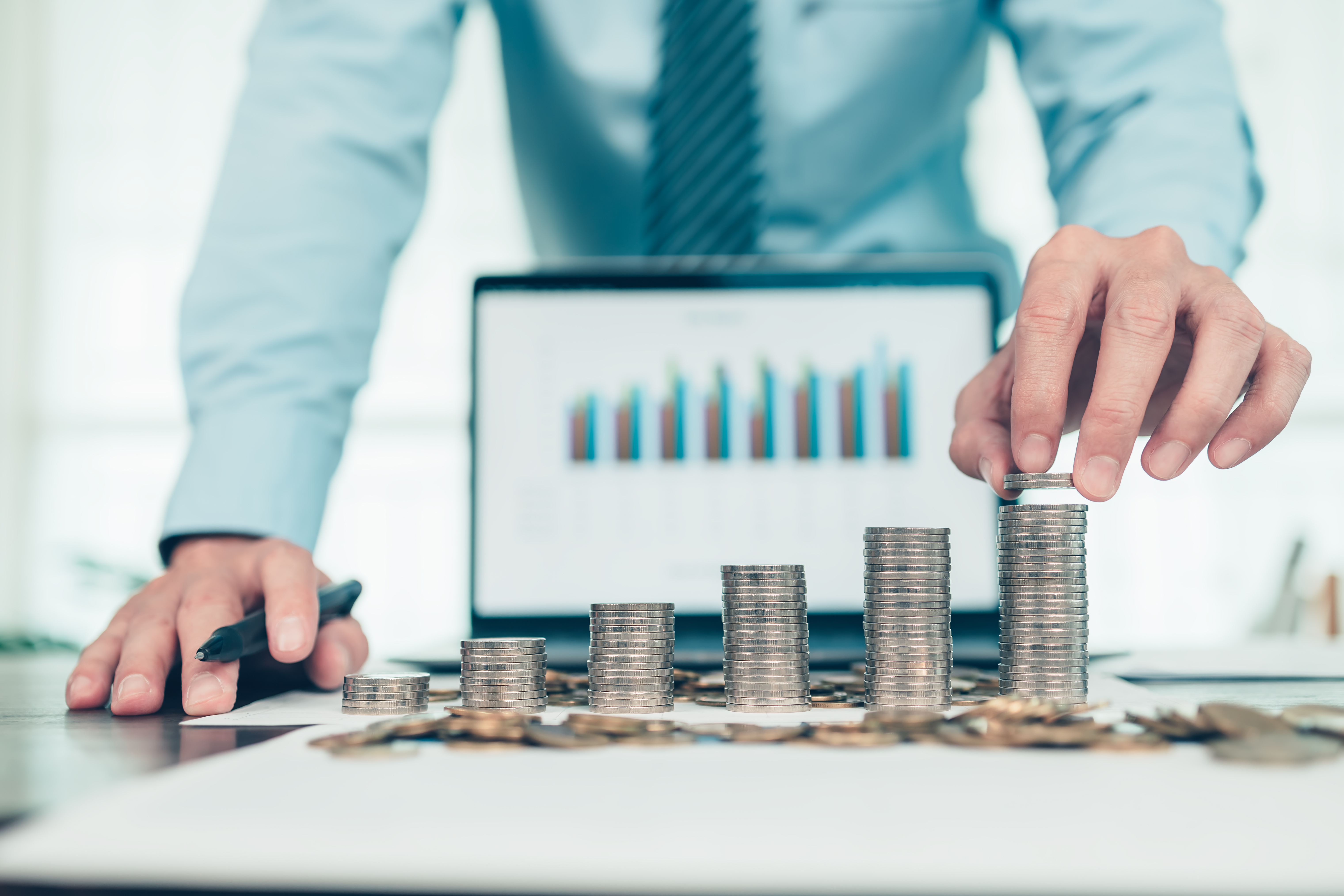A businessperson stacking coins into increasing piles on a desk with a laptop displaying a bar chart in the background, symbolizing financial growth and analysis.