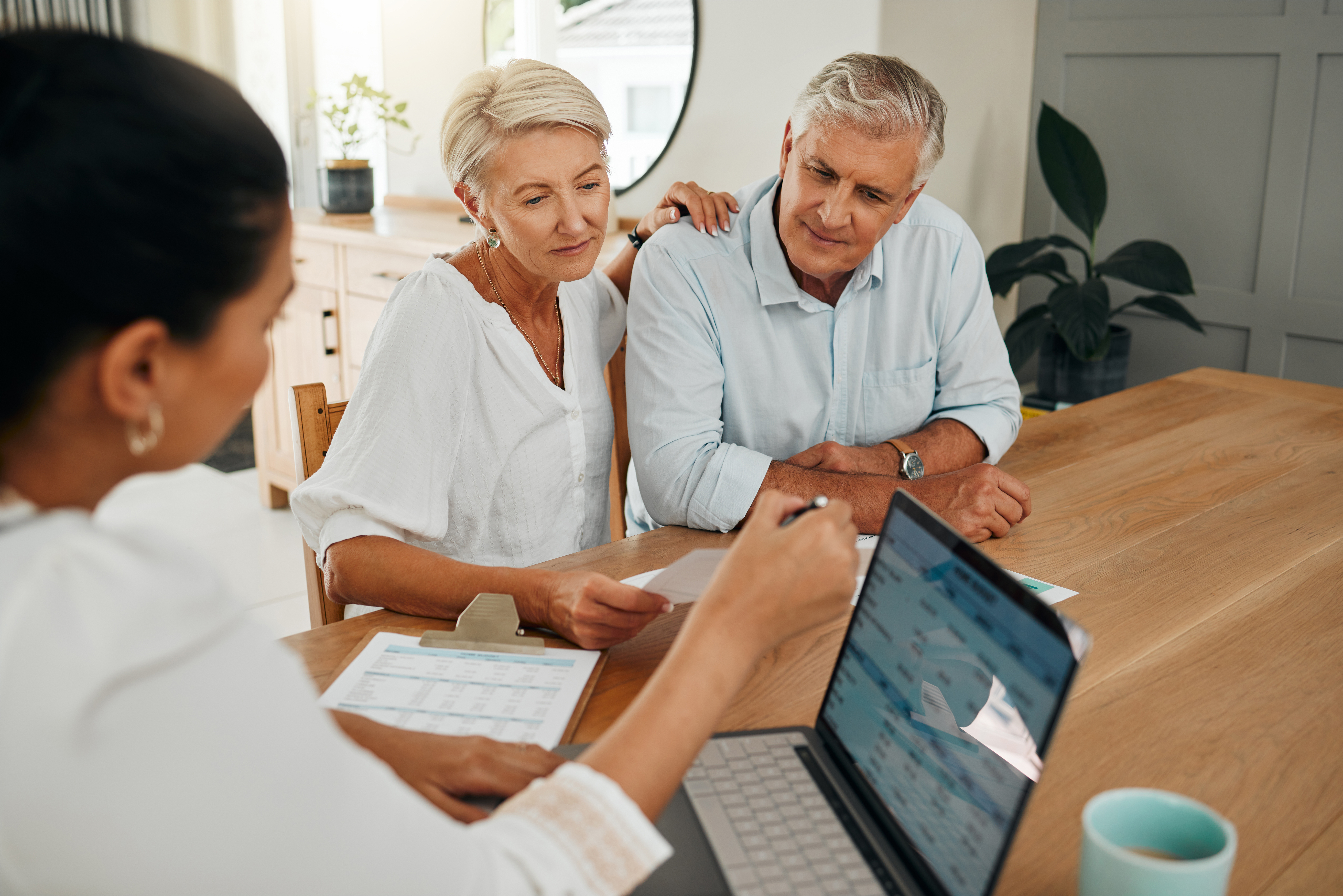 An elderly couple sitting at a table and discussing documents with a professional advisor, who is showing information on a laptop and a clipboard.