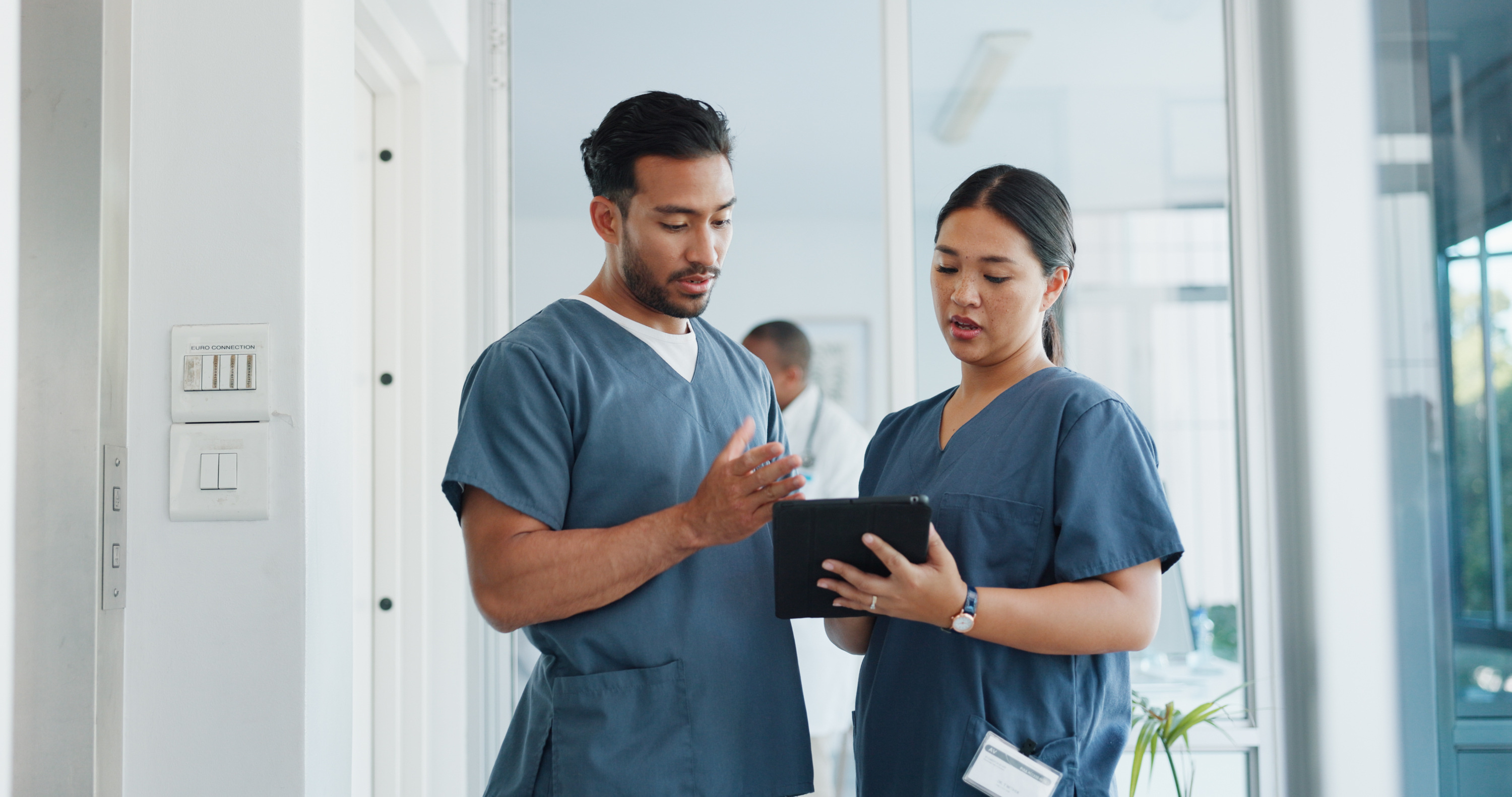 Two healthcare professionals in scrubs reviewing information on a tablet in a hospital setting, collaborating on patient care.