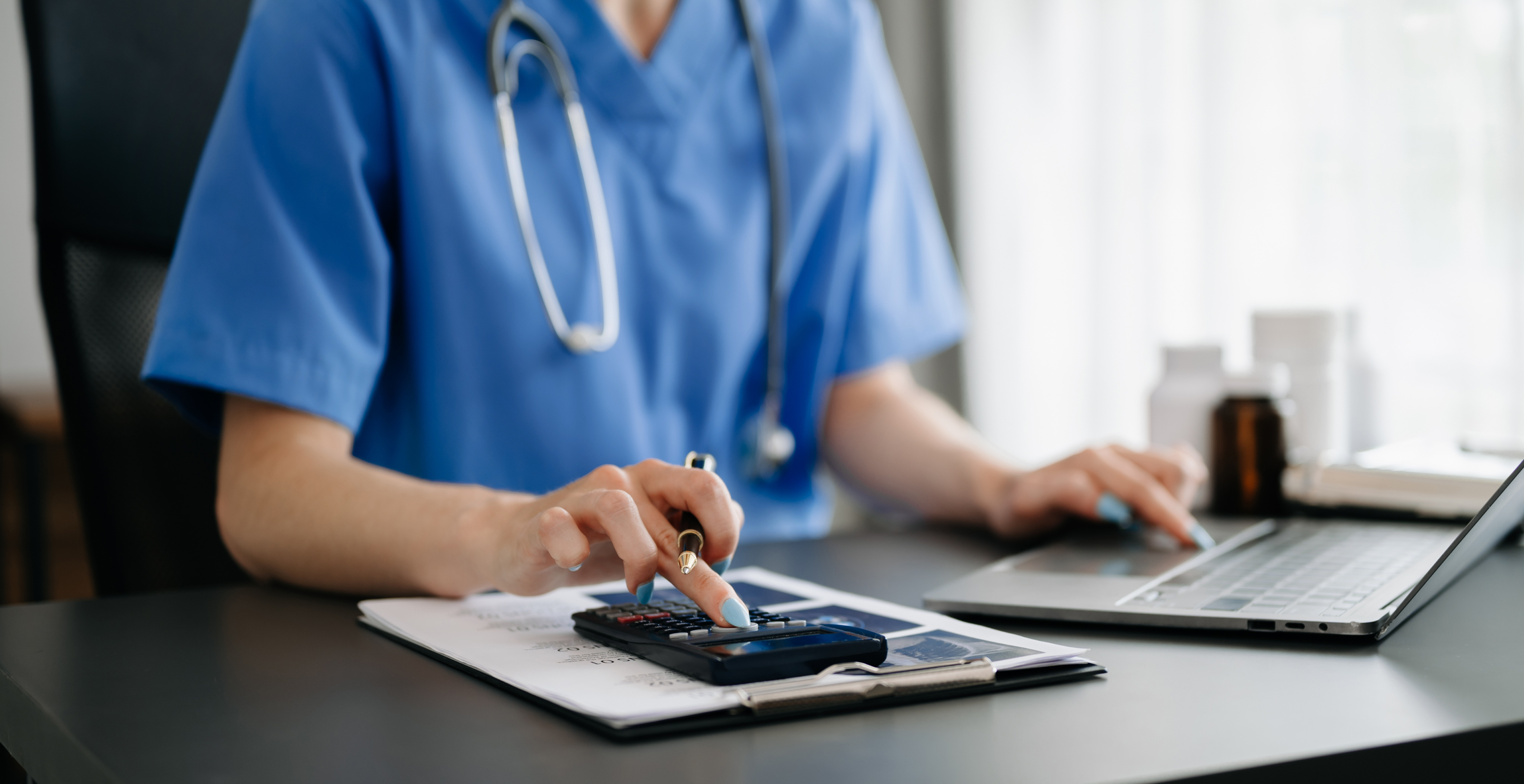 doctor sits at desk and counts on calculator while simultaneously entering results into computer. symbolizes financial organization of medical professionals.