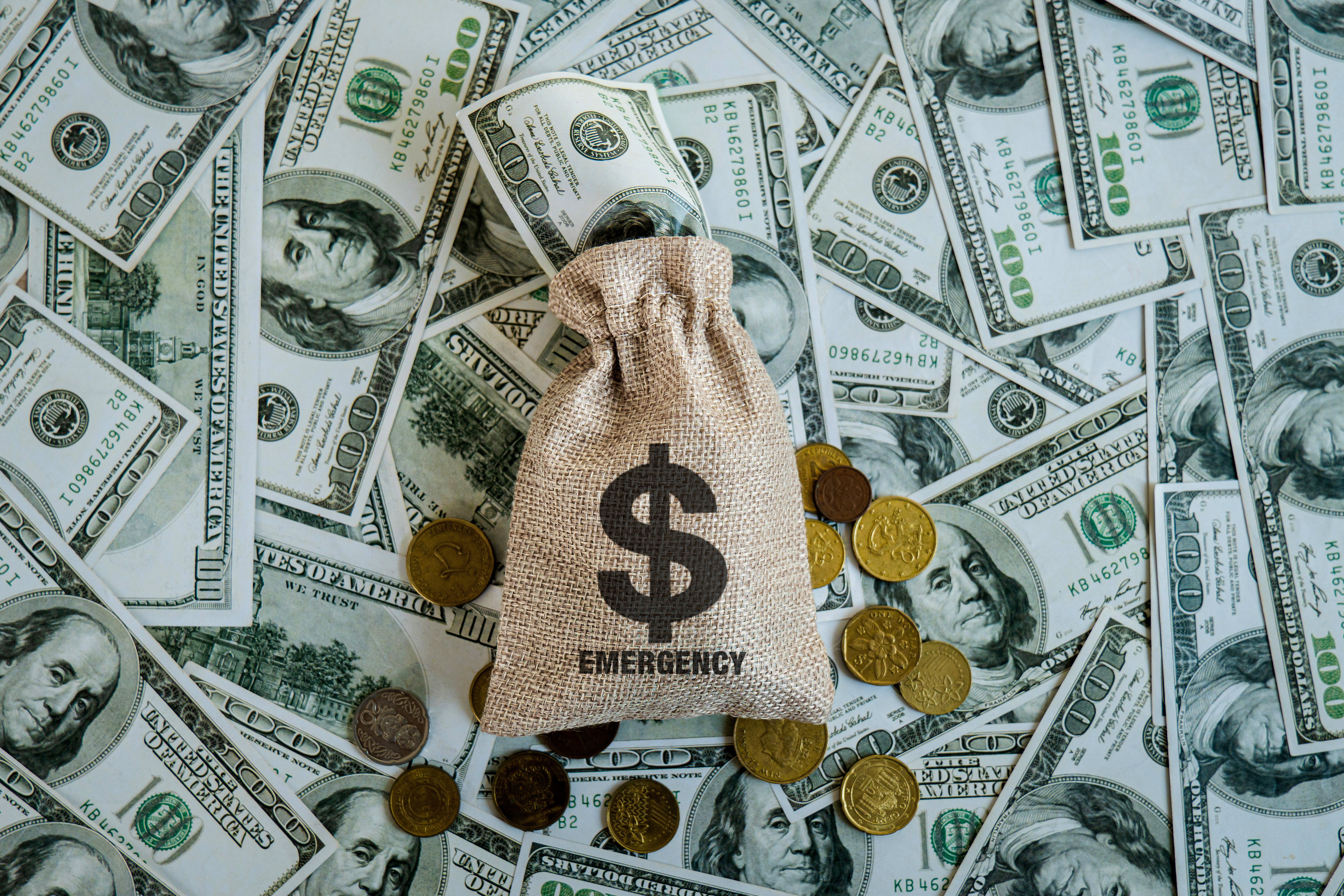 A burlap sack labeled "EMERGENCY" with a dollar sign symbol, placed on a background of scattered US dollar bills and various coins.