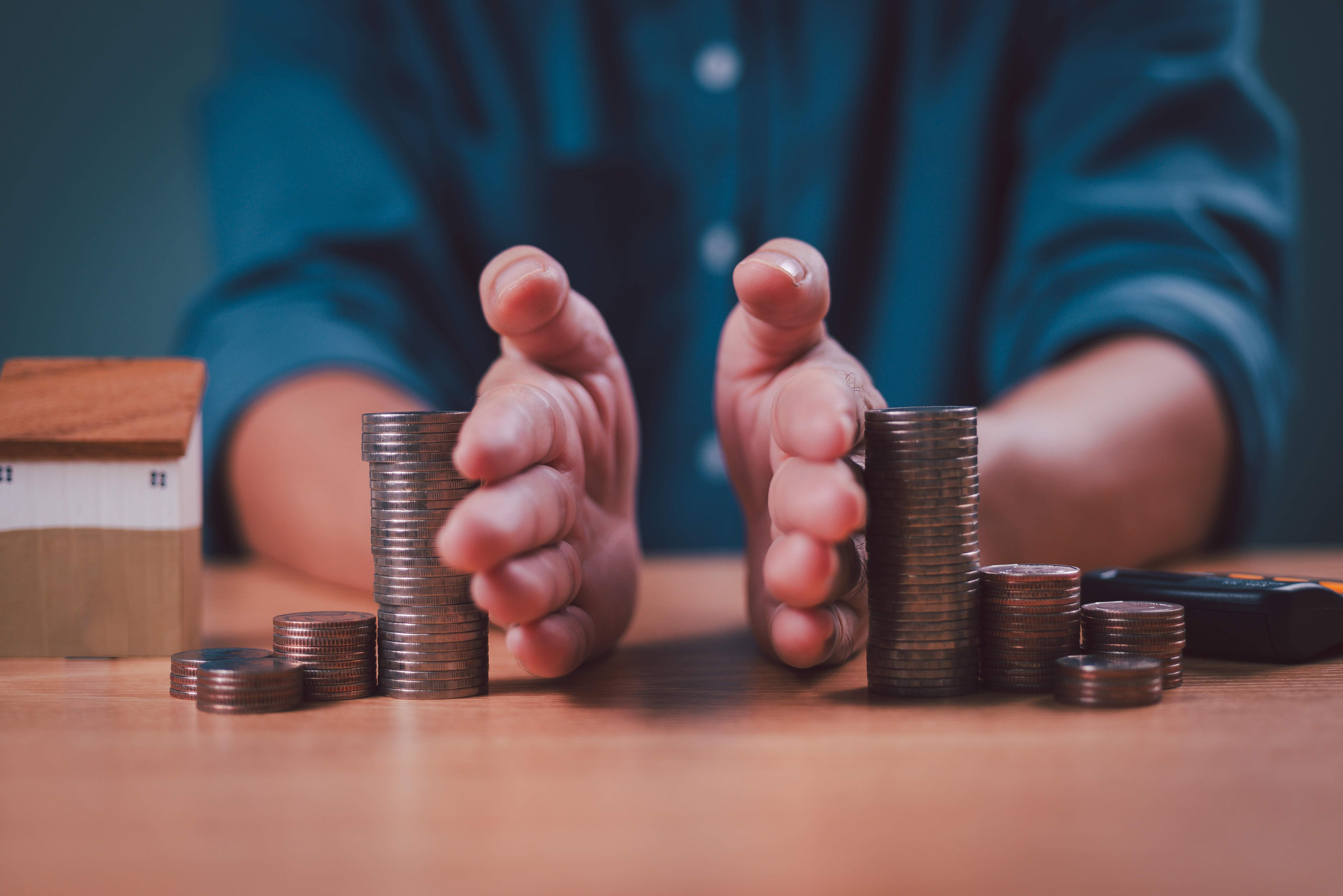 Hands organizing stacks of coins on a table next to a small house model, symbolizing financial planning and budgeting.